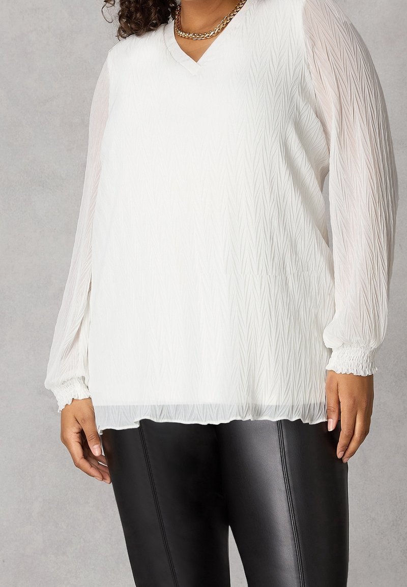 Donna che indossa una blusa bianca a maniche lunghe texturizzata con maniche trasparenti e pantaloni in pelle nera attillati, su uno sfondo neutro.