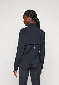 Frau mit geflochtenen Haaren in einem Pferdeschwanz, die ein schwarzes Langarmoberteil und schwarze Leggings trägt und mit dem Rücken zur Kamera steht.