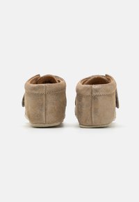 Zapatillas de ante marrón con una textura suave, que presentan una forma redondeada y un diseño sencillo. Los detalles notables incluyen adornos cosidos y una suela flexible.