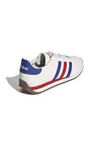 Zapatilla blanca con rayas azules y rojas, lengüeta azul en el talón, cordones blancos y suela marrón, en ángulo para mostrar el lado exterior y la parte trasera.