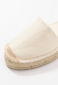Espadrille en toile beige avec un bout arrondi, semelle en corde de jute et détails cousus le long du bord. Texture lisse avec une finition naturelle.