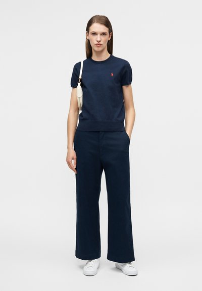 Στενομένη μπλούζα σε ναυτικό μπλε με κοντά μανίκια και ριμπ τελείωμα, συνδυασμένη με πλατύτερο παντελόνι σε ναυτικό μπλε. Το μοντέλο κρατάει μια μικρή λευκή τσάντα ώμου και φορά λευκά αθλητικά παπούτσια.