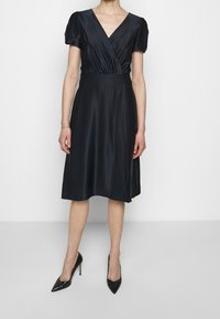 Žena v černých saténových šatech ke kolenům s krátkými nabíranými rukávy a výstřihem do V, doplněná černými špičatými lodičkami na vysokém podpatku.