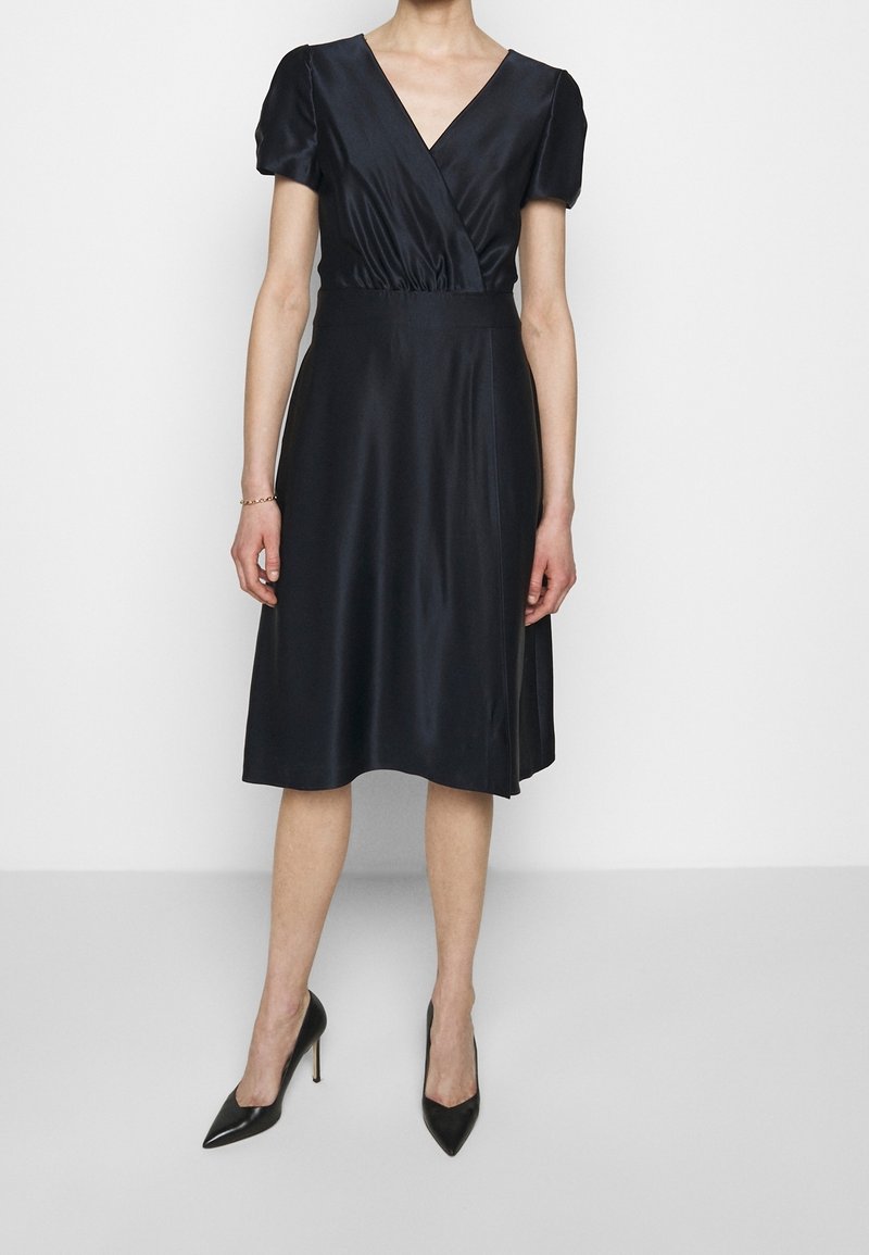 Žena v černých saténových šatech ke kolenům s krátkými nabíranými rukávy a výstřihem do V, doplněná černými špičatými lodičkami na vysokém podpatku.