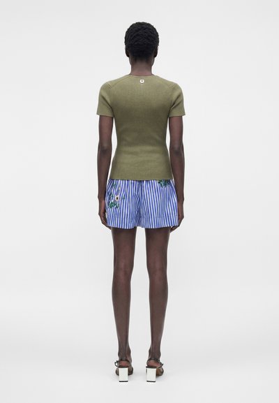 Γυναίκα που στέκεται με την πλάτη στην κάμερα, φοράει πλεκτό τοπ ελιάς με κοντά μανίκια, μπλε και άσπρο ριγέ σορτς με κεντήματα λουλουδιών και άσπρες ψηλοτάκουνες.