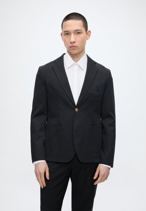 Ung mand iført en sort blazer med et våbenskjold på brystlommen over en hvid skjorte med krave, stående mod en ensfarvet hvid baggrund.