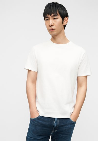 Hvid T-shirt med rund hals i bomuld. Korte ærmer, afslappet pasform. Båret med mørkeblå jeans, hænderne i lommerne, foran en ensfarvet baggrund.