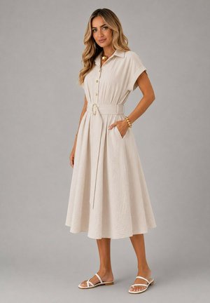 Femme portant une robe midi beige boutonnée avec une large ceinture, des manches courtes, des poches latérales, un bracelet et un collier dorés, ainsi que des sandales blanches.