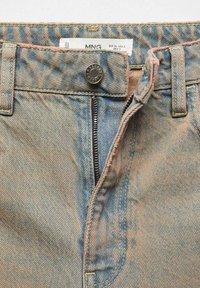 Gros plan sur le devant d'un jean en denim bleu avec une fermeture éclair partiellement défaite, un bouton en métal fermé, et une étiquette de taille visible à l'intérieur de la ceinture.