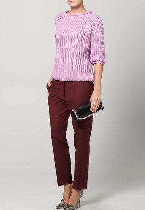 Kobieta ubrana w lawendowy sweter dziany, burgundowe spodnie o fakturze, szare szpilki, trzyma czarną kopertówkę z ozdobnym wykończeniem z cyrkonii.