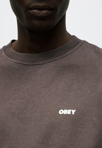 Κοντινό πλάνο ενός ατόμου που φοράει καφέ μπλούζα με στρογγυλή λαιμόκοψη και το λευκό κείμενο "OBEY" στο στήθος.