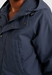 Chaqueta con capucha de color azul marino fabricada en una tela texturizada. Cuenta con botones a presión, un cierre con cremallera y un bolsillo en el lado izquierdo.