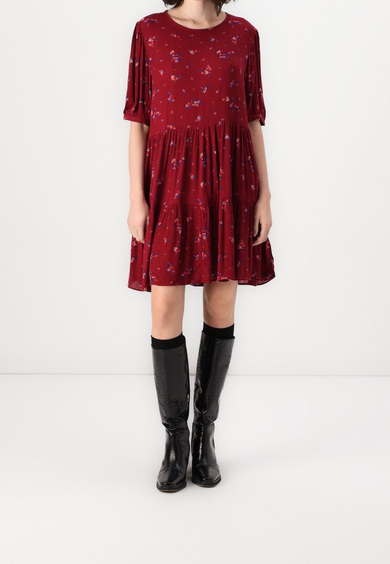Femme portant une robe rouge à fleurs fluide jusqu'aux genoux avec des manches bouffantes et des bottes noires brillantes montant jusqu'aux genoux, debout devant un fond blanc uni.