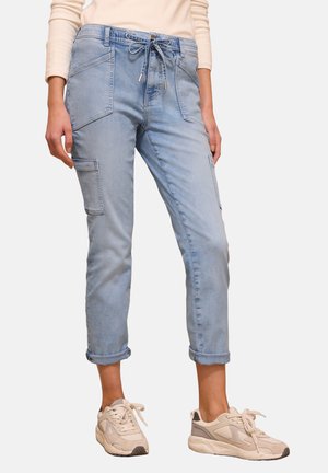 Osoba w jasnoniebieskich, mankietowanych spodniach typu cargo z przednimi kieszeniami i sznurkiem w pasie, w zestawie z beżowymi trampkami i kremową bluzką z długim rękawem.