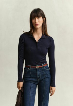 Femme aux longs cheveux bruns portant un t-shirt côtelé bleu marine à manches longues, un jean foncé taille haute et tenant une veste marron.