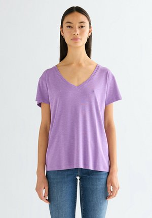 Donna con lunghi capelli scuri, che indossa orecchini a cerchio dorati, una maglietta lavanda a maniche corte con scollo a V e jeans blu, in piedi davanti a uno sfondo bianco uniforme.