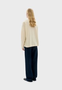 Lichtbeige overhemd met lange mouwen, losse pasvorm en verlaagde schouders, gecombineerd met donkerblauwe wijde broek en zwarte stoffen schoenen.