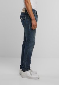 Jeans de mezclilla azul oscuro con corte recto, que presentan un diseño clásico de cinco bolsillos y acentos de costura. Combinados con zapatillas blancas.