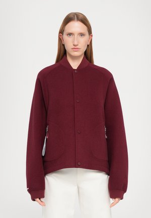 Veste bomber bordeaux avec col côtelé, dotée d'une fermeture à boutons-pression et de poches latérales avec fermetures éclair. Tissu doux et texturé.