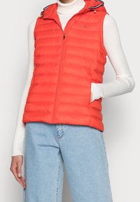 Rød puffervest med zip-front, hætte og ribbede detaljer i kanterne. Har et logo på brystet og sidelommer.