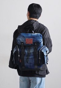 Blauwe denim rugzak met meerdere ritsen, verstelbare schouderbanden en een leren accent. Draagt op de rug van een persoon in casual kleding.