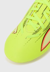 Scarpetta da calcio di un giallo brillante con una texture liscia, caratterizzata da motivi a strisce nere e accenti arancione neon. I lacci sono piatti e verdi.