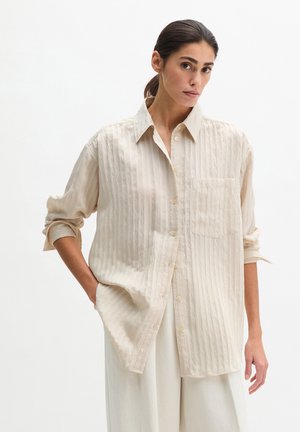 Femme portant une chemise ample texturée beige clair boutonnée avec les manches retroussées et un pantalon blanc à jambes larges, debout devant un fond uni.
