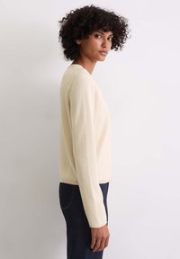 Beige langärmliger Strickpullover mit rundem Halsausschnitt und gerippten Bündchen, kombiniert mit dunklen Jeans, seitlich gezeigt.