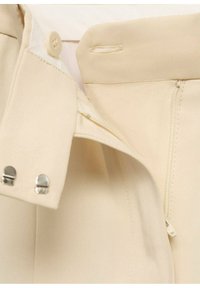 Beige-huopa, jossa on sileä rakenne, napilla ja vetoketjulla suljettuna. Viimeistelty metallisilla yksityiskohdilla vyötäröllä.