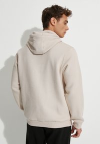 Sudadera con capucha beige de textura suave, con puños y dobladillo acanalados. Presenta una vista trasera que muestra un diseño simple, sin patrones ni logotipos.