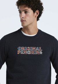 Sweatshirt marine avec un col ras du cou et un texte brodé "ORIGINAL PENGUIN" multicolore accompagné d'un graphique de pingouin sur le côté.
