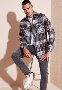 Cămașă din flanel în carouri gri și roșii, cu două buzunare pe piept, purtată peste un tricou alb, asortată cu blugi gri și adidași albi.
