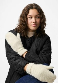 Mittens color crema con una texture a coste e orli risvoltati, realizzati in un tessuto morbido, indossati sopra un giubbotto trapuntato nero e una maglietta beige.