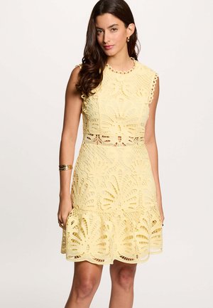 Donna con capelli lunghi e scuri indossa un vestito giallo in pizzo senza maniche con ritagli floreali e orlo smerlato, accessoriato con bracciale e orecchini d'oro.
