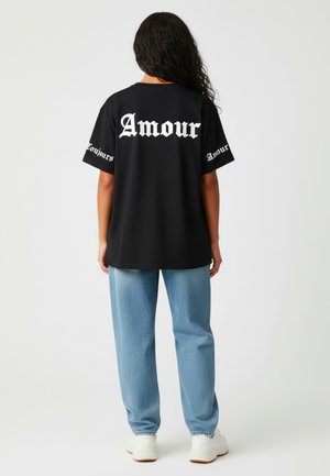 Zwart oversized t-shirt met witte gotische tekst "Amour" op de achterkant en mouwen, gecombineerd met lichtblauwe patroonbroek en witte sneakers.