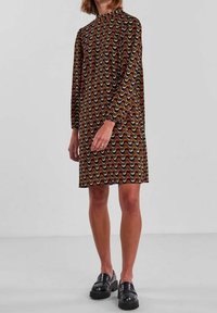 Brun klänning med lång ärm, hög halsringning och vågmönster i kräm, orange och grön. Bärs med svarta kraftiga loafers.