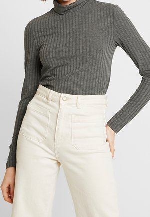 Kobieta ubrana w dopasowany, szary, prążkowany, długi rękaw wsunięty w wysokie, szerokie spodnie w kolorze kremowym z naszywanymi kieszeniami z przodu.