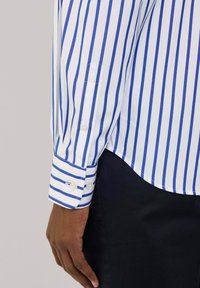 Camisa blanca con rayas verticales azules, con puños abotonados y un dobladillo curvado. Tela suave, diseño estructurado y líneas limpias.
