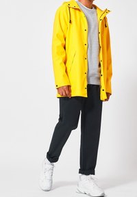 Gelbe wasserdichte Jacke mit Kapuze, Druckknöpfen, frontalen Taschen, getragen über einem grauen Sweatshirt und schwarzen Hosen, kombiniert mit weißen Sneakers.