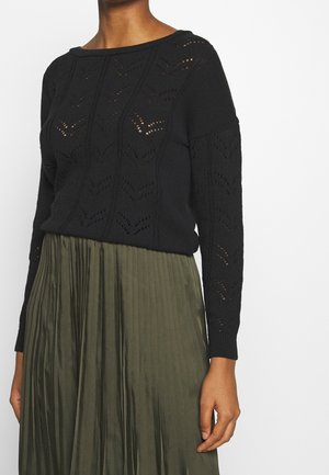 Frau trägt einen schwarzen, langärmligen Strickpullover mit Chevron-Ausschnitten und einen olivgrünen plissierten Rock vor einem schlichten Hintergrund.