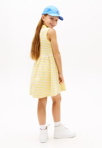 Κορίτσι με χαμόγελο και μακριά μαλλιά, που φοράει κίτρινο και λευκό ριγέ φόρεμα, μπλε καπέλο, λευκές κάλτσες και λευκά αθλητικά παπούτσια, στέκεται μπροστά σε λευκό φόντο.