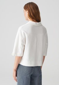 Weißes Kurzarmoberteil mit Kragen und strukturiertem Stoff. Die Rückseite hat eine Naht. Getragen mit blau-weißen gestreiften Hosen.