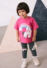 Camiseta de color rosa brillante con un diseño de unicornio y arcoíris, combinada con leggings grises que presentan motivos similares y detalles blancos con volantes.