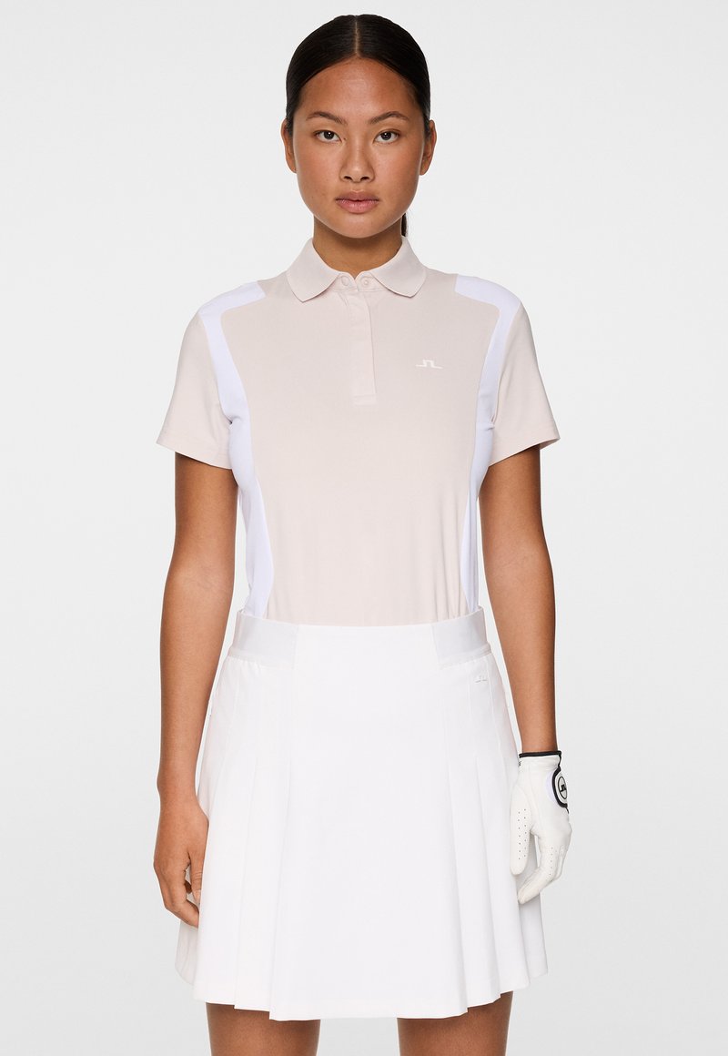 Kobieta ubrana w beżową i białą koszulę golfową, białą plisowaną spódnicę oraz białą rękawicę golfową na prawej ręce, stojąca na tle jednolitym.