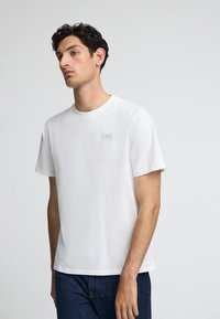 Witte katoenen T-shirt met korte mouwen, ronde hals en een klein monochroom logo op de borst. Gecombineerd met blauwe spijkerbroek.