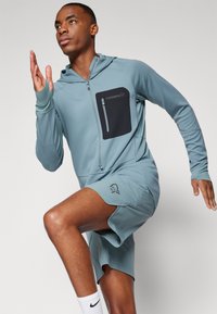 Un sweat à capuche de sport bleu clair avec une poche poitrine zippée noire, des manches longues et un short assorti, arborant un logo sur le short.