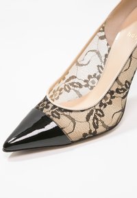 Chaussure à talon haut à bout pointu avec embout en cuir verni noir et corps en dentelle transparente ornée de motifs floraux.