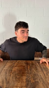 Oseba s kratkimi temnimi lasmi nosi črno majico, sedi za lesenim mizo z teksturirano površino in svetlo obarvano steno v ozadju.