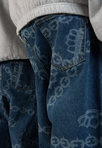 Vaqueros de mezclilla con un lavado medio, que presentan un patrón gráfico repetido de cadenas y puños, bolsillos cosidos y detalles de costura en contraste.