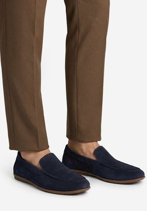 Mocassini slip-on in camoscio blu navy indossati con pantaloni marroni su misura, che mostrano dettagli laterali e superiori della scarpa su sfondo bianco.